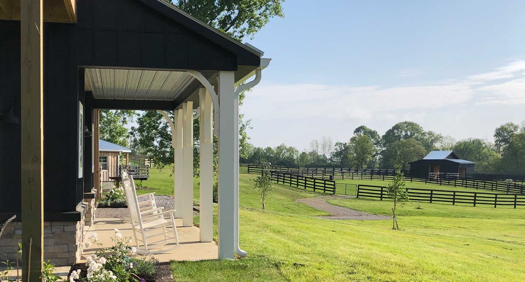 Covered porch with white rocking chairs overlooking a peaceful horse farm with green paddocks and black farm fencing
